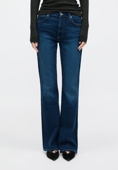 Γυναίκα που φοράει σκούρα μπλε τζιν με φαρδιά πόδια, μαύρες γόβες με μυτερά άκρα και σκούρο μακρυμάνικο τοπ σε απλό λευκό φόντο.