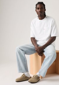 Λευκό βαμβακερό μπλουζάκι με τσέπη μπροστά, ανοιχτό μπλε τζιν παντελόνι, μπεζ ξύλινα παπούτσια τύπου κλογκ, και λευκές κάλτσες, καθισμένος σε ξύλινο τεμάχιο.