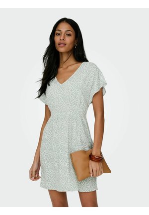 Kvinne iført en lys grønn kort kjole med mønster og V-hals, holder en beige clutch og har et brunt armbånd på høyre håndledd.