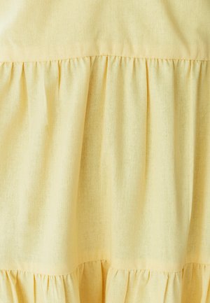 Gaiši dzeltenas krāsas kleita ar teksturētu audumu un volānu kārtām, kas izceļ mīkstu un gaisīgu dizainu.