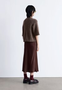 Braun-flauschiger Strickpullover mit kurzen Ärmeln, kombiniert mit einem langen braunen SatRock, burgunderfarbenen Socken und schwarzen Schnürschuhen, von hinten betrachtet.