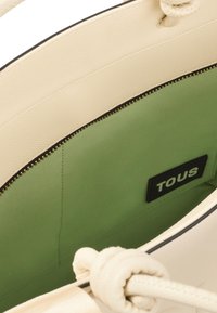 Beżowa torba na ramię z teksturowanym wykończeniem, zieloną podszewką i zapięciem na zamek. Posiada czarną metkę "TOUS" oraz węzłowe uchwyty.
