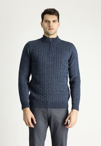 Marineblauer Strickpullover mit hohem Reißverschlusskragen, der ein Zopfmuster sowie einen gerippten Saum und Ärmelbündchen aufweist. Das Modell trägt ihn mit grauen Hosen.