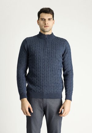 Marineblauwe gebreide trui met een hoge ritskraag, voorzien van een kabelbreipatroon en ribbelboorden aan de onderkant en mouwen. Model draagt het met grijze broek.