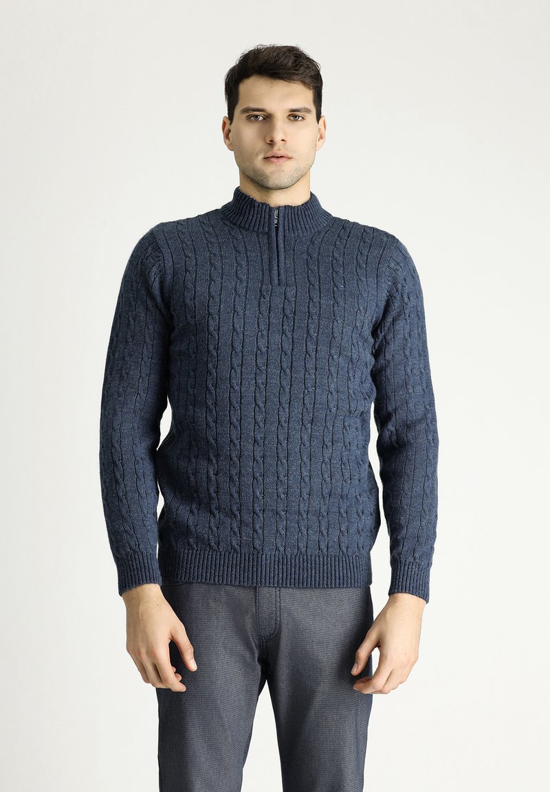Marineblauer Strickpullover mit hohem Reißverschlusskragen, der ein Zopfmuster sowie einen gerippten Saum und Ärmelbündchen aufweist. Das Modell trägt ihn mit grauen Hosen.