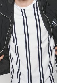 Svart jacka med dragkedja över en vit skjorta med vertikala marinblå ränder. Skjortan har en rund hals och en slät yta; jackan har en logotypdetalj.