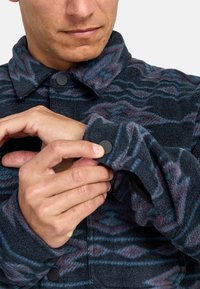 Giacca in pile con un motivo a onde nei colori blu scuro, viola e grigio. Presenta un colletto da camicia e un bottone a pressione nero ai polsini.