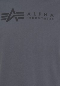 Tamsiai mėlynas medvilninis marškinėliai su dideliu juodu logotipu, stilizuota "A" ir sparnais virš teksto "ALPHA INDUSTRIES." Lygus paviršius, apvalus iškirpimas.