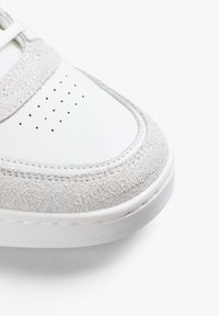 White leather sneaker with grey suede accents, round toe, perforated detailing on the toe box, and a textured rubber sole.