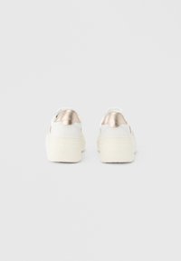 Λευκά αθλητικά παπούτσια με μεταλλικές χρυσές λεπτομέρειες στη φτέρνα, υλικό με υφή και χοντρή σόλα. Εμφανής σήμανση κοντά στη φτέρνα.