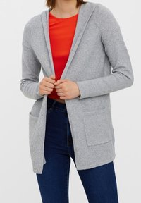 Ljusgrå cardigan med huva, med två framfickor. Tyget verkar mjukt och strukturerat, lager på lager över en röd topp.