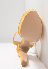 Keltainen mokkajalkine avonaisella varpaalla, hoikalla korolla ja sileällä beige ulkopohjalla. Varusteena nilkkastrappi tuen saamiseksi.