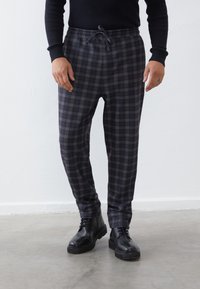 Un pantalon de jogging à carreaux gris foncé avec une taille élastique et un cordon de serrage, associé à des bottines noires sur un sol en béton.