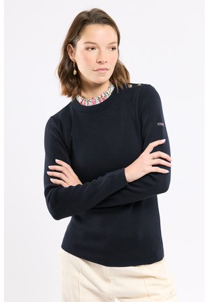 Femme aux cheveux bruns mi-longs portant un pull bleu marine sur un chemisier à col fleuri, les bras croisés, regardant légèrement sur le côté sur fond blanc.