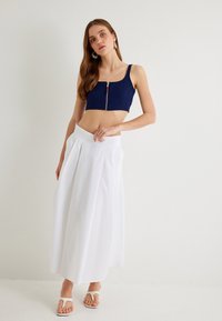 Marineblå cropped top med lynlås foran, parret med en svajende hvid maxiskirt. Skørten har høj talje og plisseret design. Skoene er hvide sandaler.