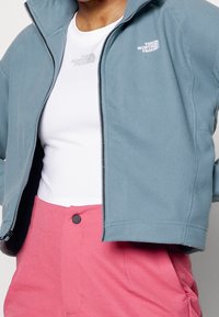 Jasnoniebieska polarowa kurtka z zamkiem błyskawicznym i wysokim kołnierzem. Posiada wyszywane logo na klatce piersiowej, zestawiona z białą koszulą i różowymi spodniami.