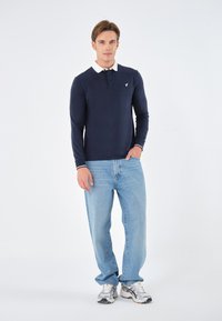Marineblaues Langarm-Polo mit weißem Kragen, gesticktem Logo auf der Brust, kombiniert mit hellblauen Jeans und silbernen Sportschuhen.