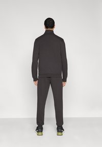 Σκούρο γκρι φόρμα με ψηλό γιακά, μακριά μανίκια και ελαστικές μανσέτες· ταιριαστά παντελόνια· φορεμένα με μαύρα και πράσινα αθλητικά παπούτσια.