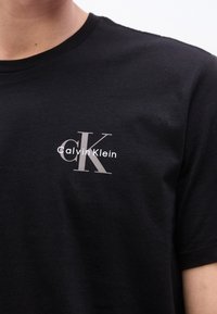 Μαύρο κοντομάνικο μπλουζάκι με λευκό και γκρι λογότυπο "Calvin Klein" τυπωμένο στο πάνω αριστερό μέρος του στήθους.