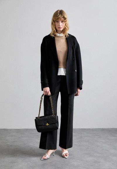 Casaco oversized preto, suéter castanho, camisa branca; calças largas pretas. Bolsa preta com alça de corrente dourada, sandálias brancas de salto. Fundo texturizado.