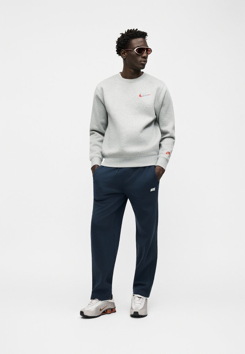 Man wearing gray Nike sweatshirt, navy Nike sweatpants, white Nike sneakers, and red-tinted sunglasses, standing with hands in pockets against white background.