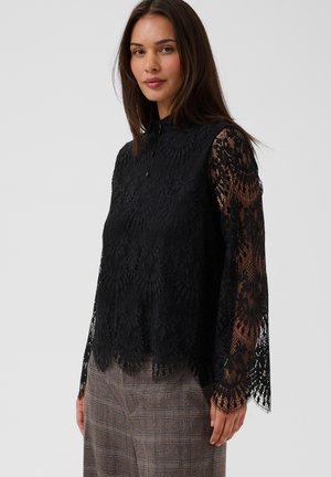 Vrouw draagt een zwarte kanten blouse met lange mouwen en bloemenpatroon en een bruine geruite broek, staand tegen een effen lichte achtergrond.