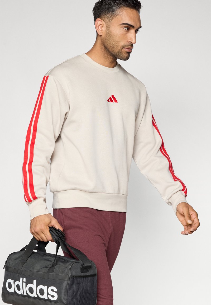 Man in beige sweatshirt met rode strepen en logo, bordeauxrode broek, met zwarte Adidas sporttas, loopt tegen een effen achtergrond.