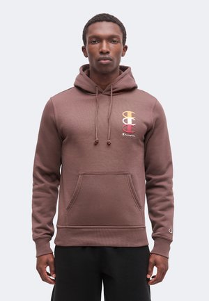 Man draagt een bruine Champion-hoodie met een kleurrijk logo op de borst en een klein logo-patch op de linkermouw, staand tegen een effen achtergrond.