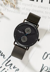 Orologio con cinturino in rete nera, quadrante nero texturizzato, due sottocampi grigi, accenti argento e dettagli blu. Presenta un design minimalista.