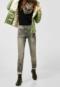 Person iført en lys grøn pufferjakke, sort top, mønstret tørklæde, falmede grå jeans med opkortning og sorte ankelstøvler mod en ensfarvet baggrund.