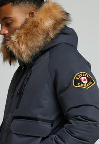 Ciemnoszara parka z sztucznym futrem na kapturze, z zamkiem błyskawicznym z przodu i kieszeniami po bokach. Posiada żółto-czarną naszywkę z napisem "Zavetti Canada".