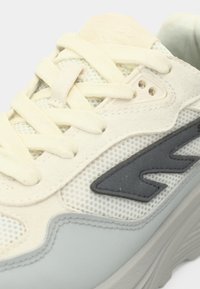 Svijetlosiva tenisica s teksturiranom mrežastom gornjom stranom, kremasto obojenim vezicama i tamnosivim detaljem logotipa sa strane.