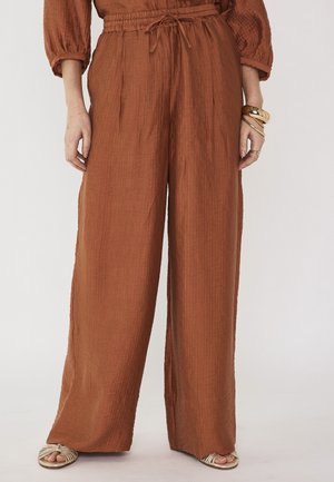 Pantalon large de couleur rouille avec taille à cordon, porté avec des sandales ouvertes beiges et des bracelets dorés au poignet gauche.