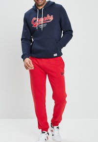 Felpa navy con laccio bianco e logo rosso, abbinata a pantaloni tuta rossi e sneaker bianche. Tessuto di cotone, vestibilità casual, tasca frontale.