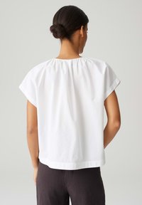 Weiße Baumwollbluse mit lockerer Passform, kurzen Ärmeln und gerafftem Halsausschnitt. Der Stoff ist glatt und leicht, mit gleichmäßiger Textur.