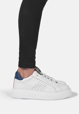 Person iført sorte leggings og hvide lædersneakers med blå hælflap og perforerede sidetaljer, stående på en hvid overflade.