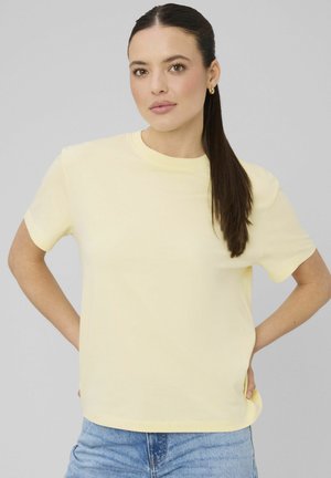 Femme aux longs cheveux bruns attachés en queue de cheval, portant de petites boucles d'oreilles en anneau doré, un T-shirt jaune clair et un jean bleu clair, debout les mains sur les hanches.