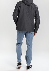 Giacca impermeabile grigio scuro da uomo con cappuccio, abbinata a jeans blu chiaro e scarpe da ginnastica nere. Tessuto liscio con dettagli minimi.