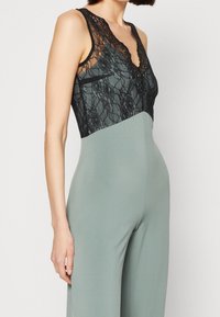 Jumpsuit bez rukávů s černou krajkovou korzetovou částí a světlým zeleným, přiléhavým spodním dílem. Tkanina je hladká s mírným leskem.