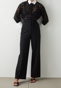 Czarna koronkowa bluzka z białym kołnierzykiem, zestawiona z wysokim stanem czarnymi spodniami z przewiązanym paskiem, podkreślonym szerokimi nogawkami. Czarne sandały.