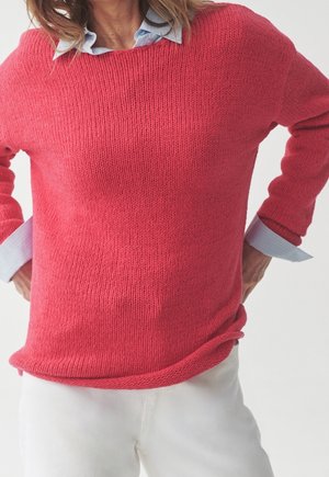 Roze gebreide trui met een brede halslijn en lange mouwen, over een lichtblauw overhemd met kraag gedragen. Witte broek zichtbaar aan de onderkant.