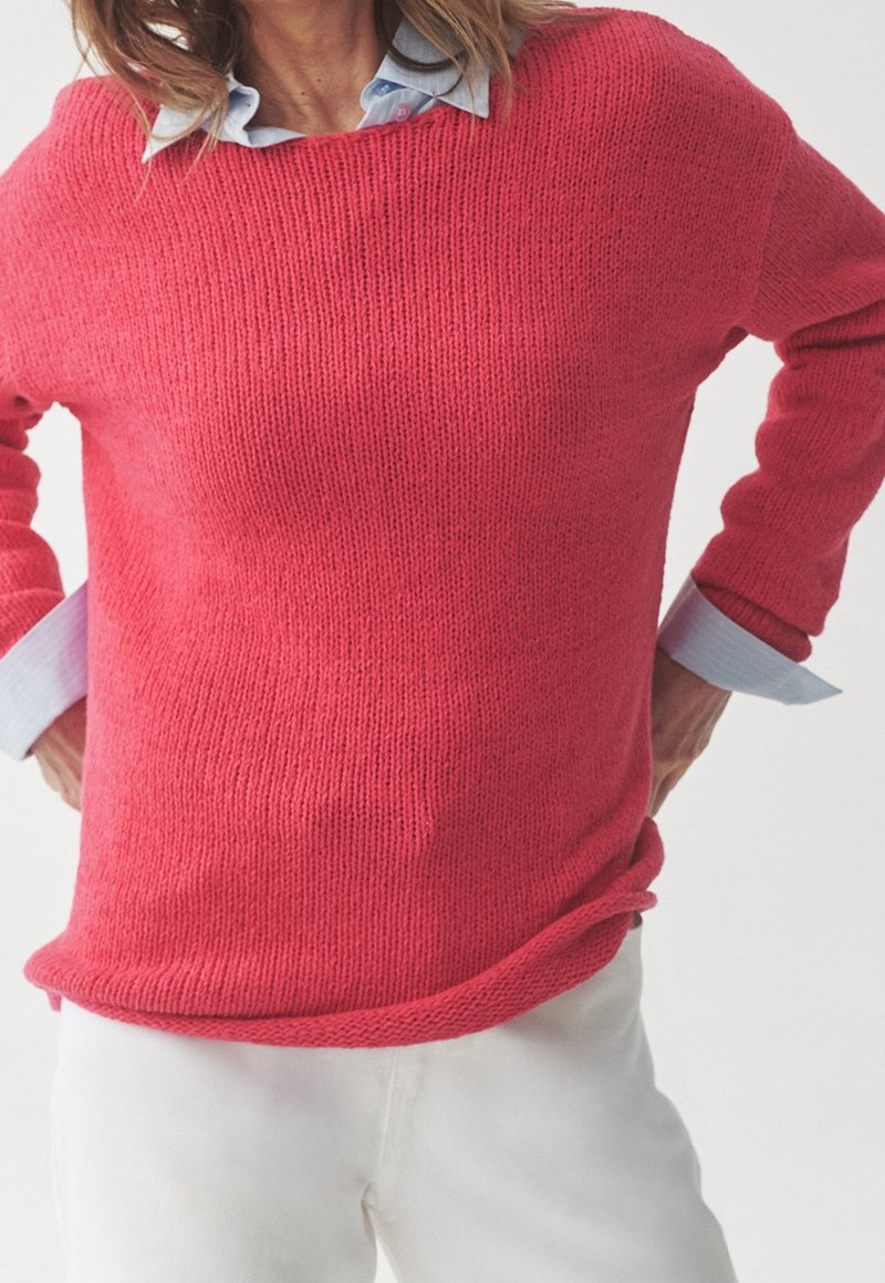 Rosa gestrickter Pullover mit weitem Ausschnitt und langen Ärmeln, über einem hellblauen Hemd mit Kragen getragen. Weiße Hosen sind am Boden sichtbar.
