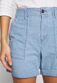 Shorts à rayures verticales bleues et blanches avec ourlets retroussés et poches plaquées avant, portés avec un haut blanc à manches longues.