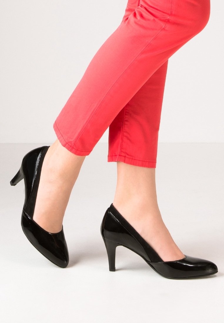 Femme portant un pantalon rouge court et des chaussures à talons moyens en cuir verni noir, debout avec un pied légèrement levé.