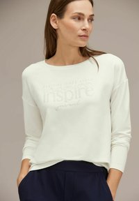 Felpa bianca con maniche lunghe e scollatura rotonda presenta il testo in rilievo "TEMPI ECCITANTI AVANTI ispira la tua mente." Abbinata a pantaloni blu navy.