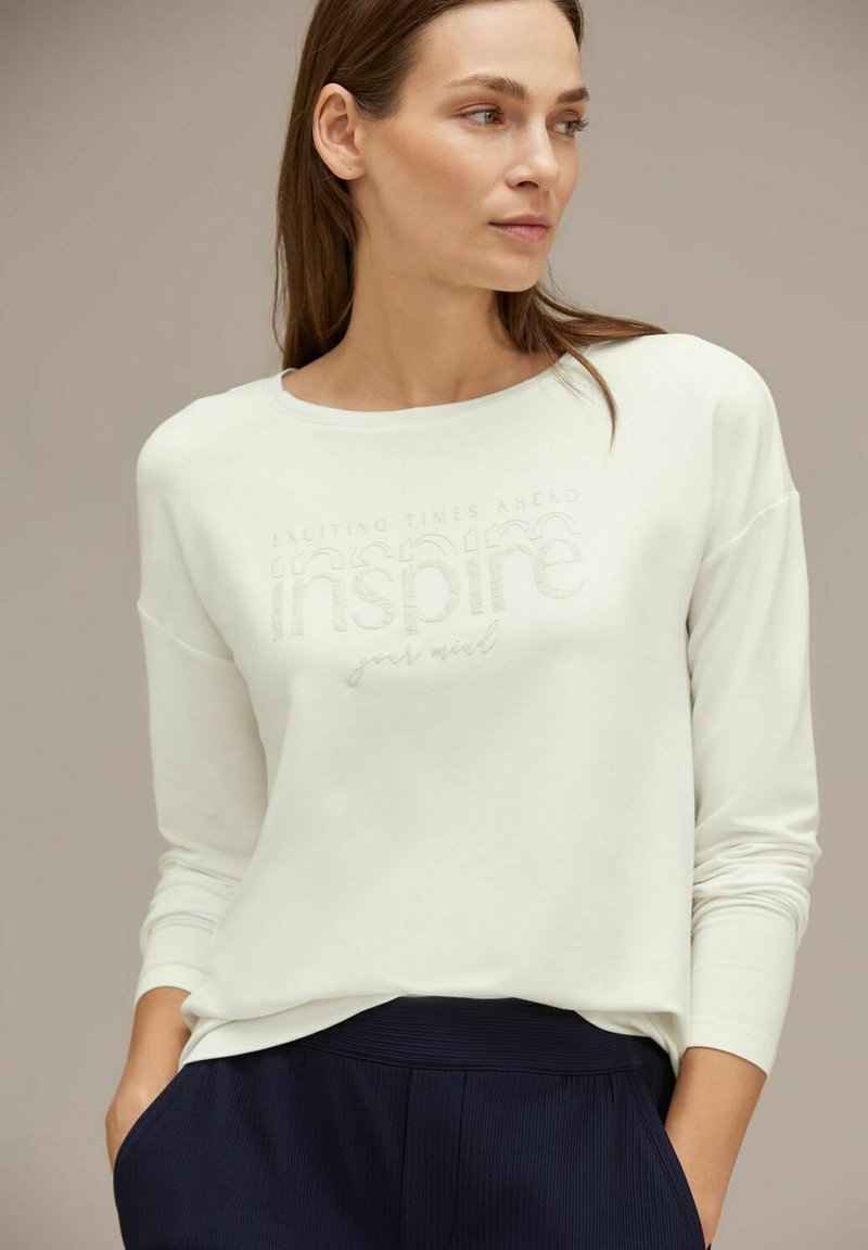 Felpa bianca con maniche lunghe e scollatura rotonda presenta il testo in rilievo "TEMPI ECCITANTI AVANTI ispira la tua mente." Abbinata a pantaloni blu navy.