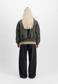 Person with long blonde hair wearing a dark green bomber jacket, beige top, and loose black pants, standing against a plain white background.