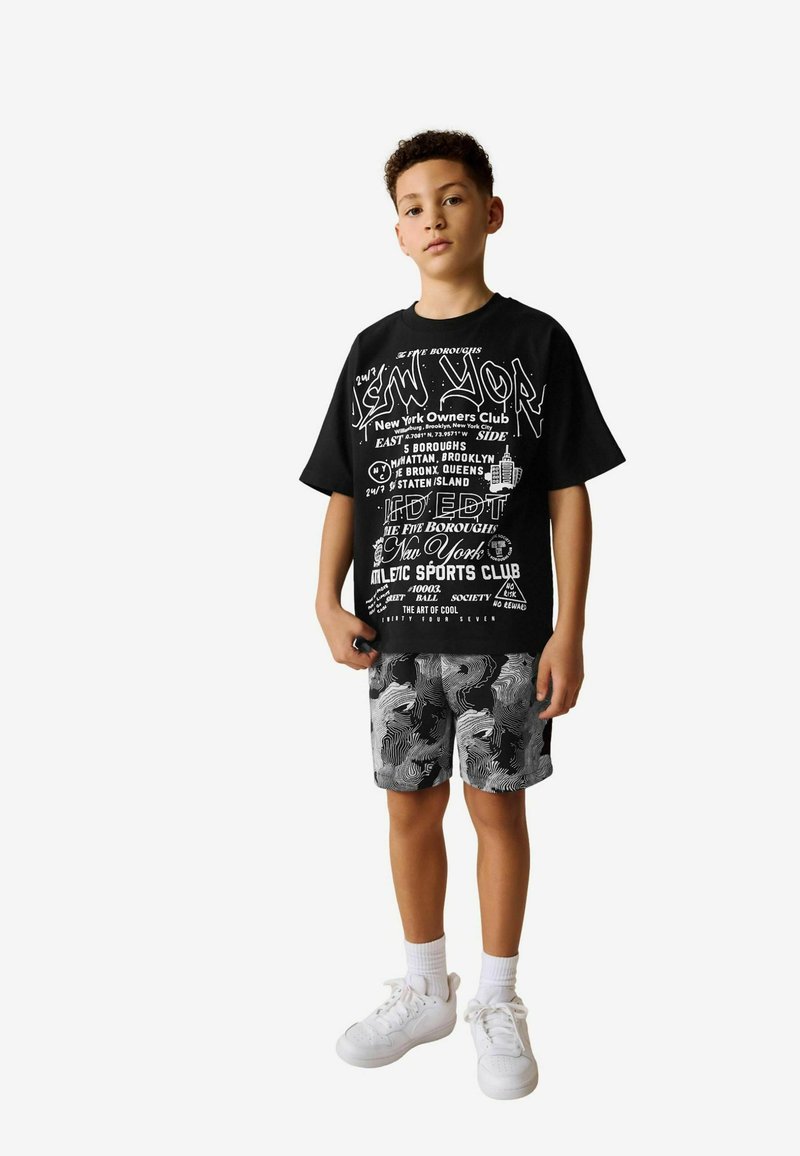 Zwart t-shirt met witte grafische tekst, gecombineerd met zwart-witte gestreepte shorts en witte sneakers met gestructureerde zolen.