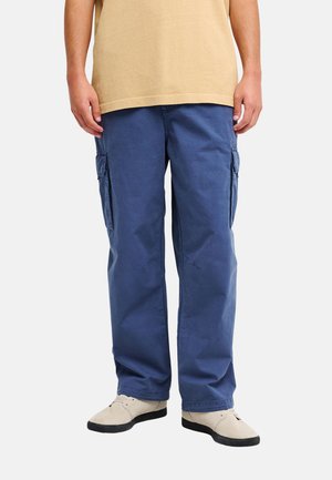 Osoba ubrana w niebieskie spodnie cargo, beżowy t-shirt i jasnobeżowe buty stoi na tle jednolitego tła.