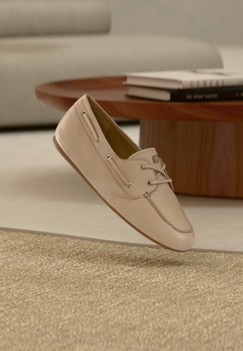 Mocasini din piele bej cu șireturi și detalii de cusătură, plutind deasupra unui covor bej țesut într-un decor modern de living.